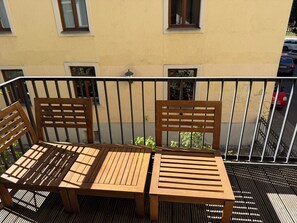 A wooden table and chairs on a balcony with a metal railing, a yellow building in the background, and a red car parked on the street. - Modern, Fully Furnished City Apartment - 5 minutes from downtown (München)