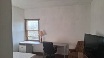 A bedroom with a single bed, a desk with a chair, a television, and a window with blinds.