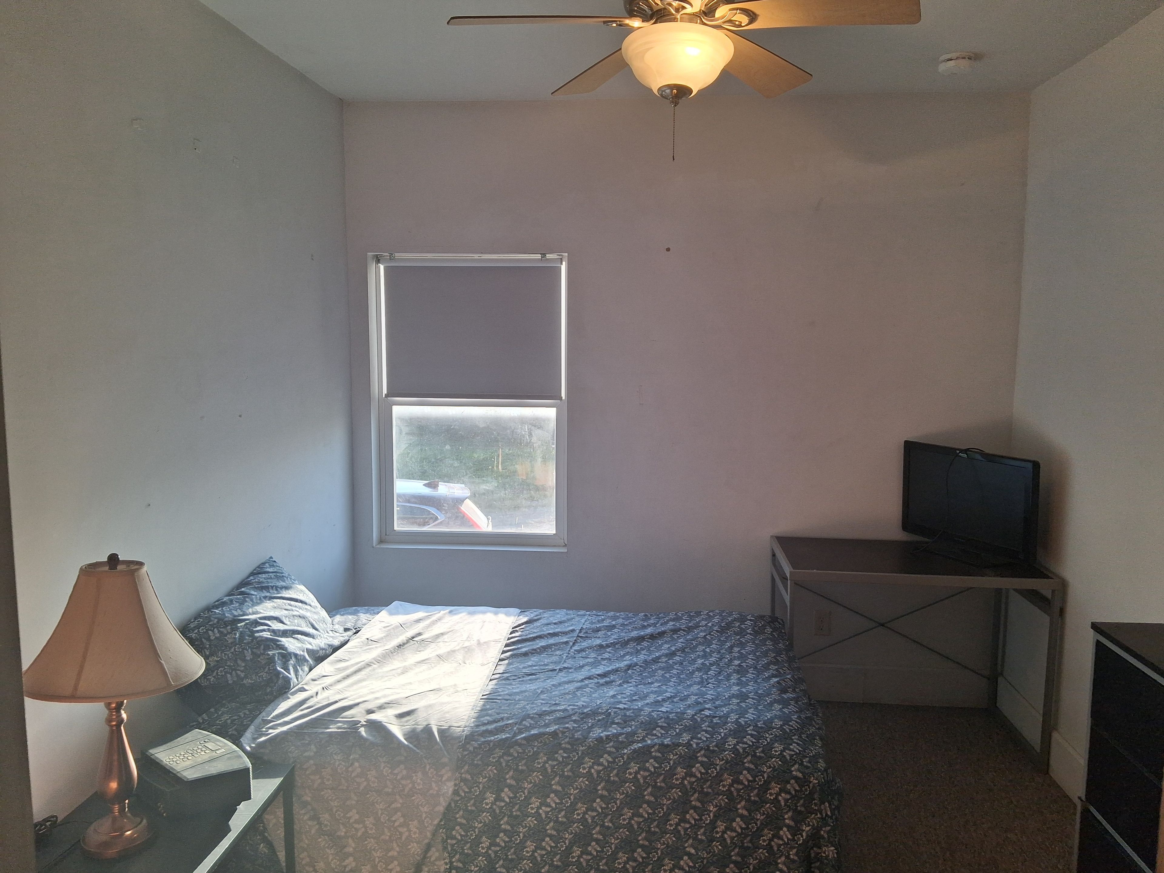 A small bedroom with a bed, a desk with a television, a window, and a ceiling fan.