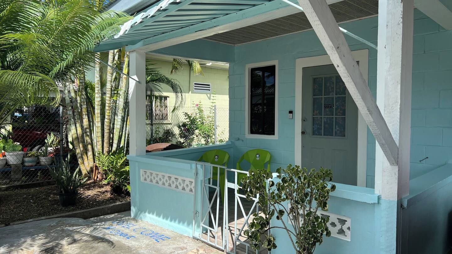 A small, light blue building with a covered porch, two green chairs, and a potted plant.