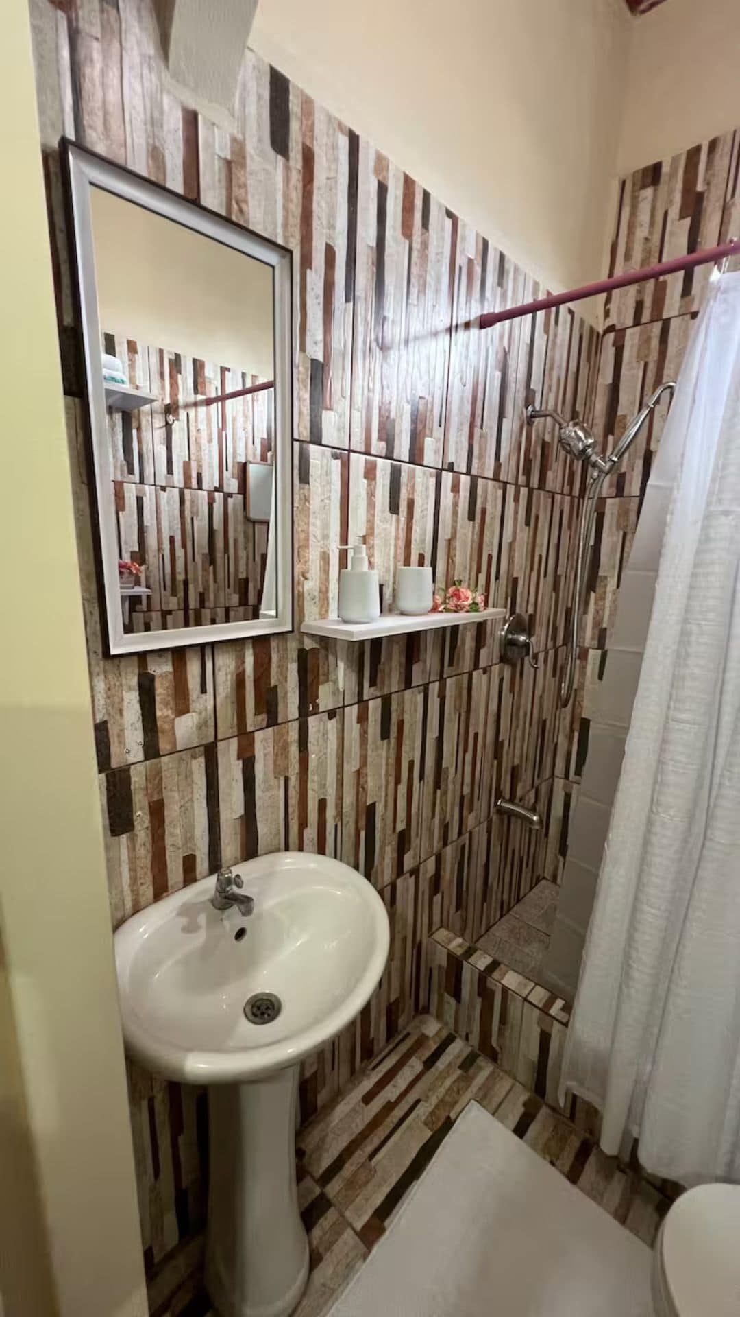 A bathroom with a sink, mirror, shower, and a wall with a vertical wood pattern.