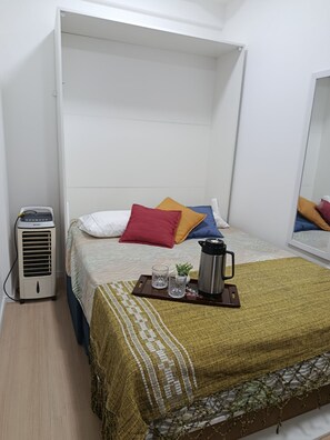 A single bed with a white headboard, a small bedside table with a thermos, glasses, and a plant, and a standing heater. - New Studio República Copan- Mackenzie (São Paulo)
