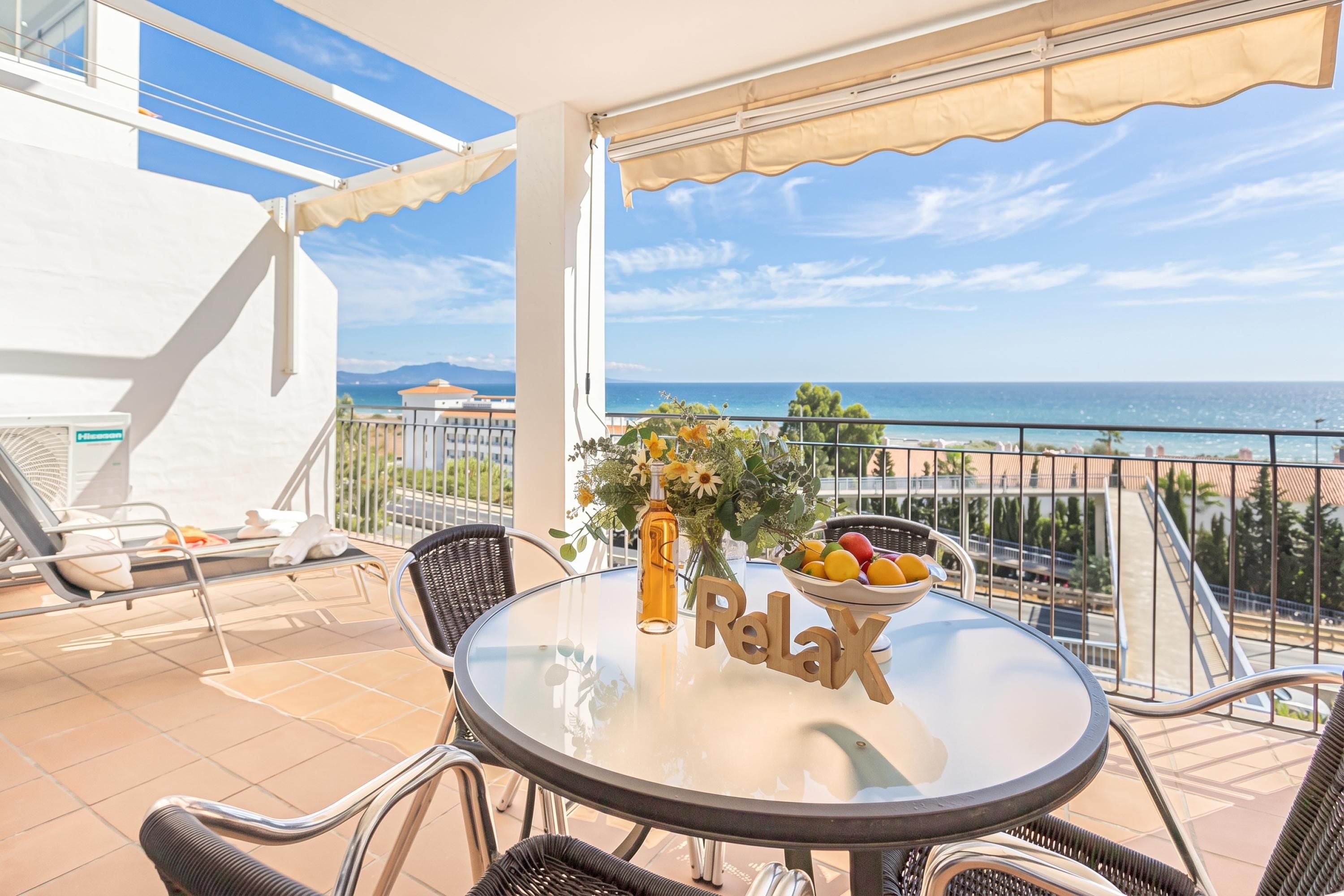 A balcony with a glass table, wicker chairs, a bottle, and a bowl of fruit.