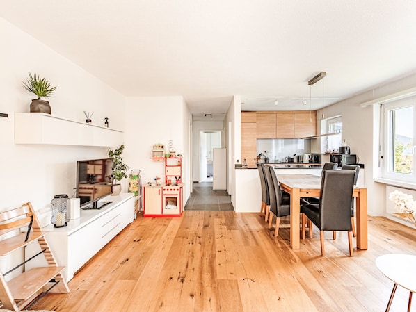 A modern kitchen with wooden cabinets, a dining area with a table and chairs, a TV on a white cabinet, and a hallway leading to another room.