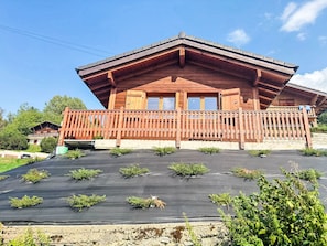 A wooden chalet with a balcony, a flat roof, and a garden area with plants.