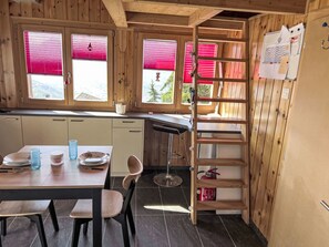 Een kleine keuken met houten wanden, een eethoek met tafel en stoelen, en een raam met rode jaloezieën.