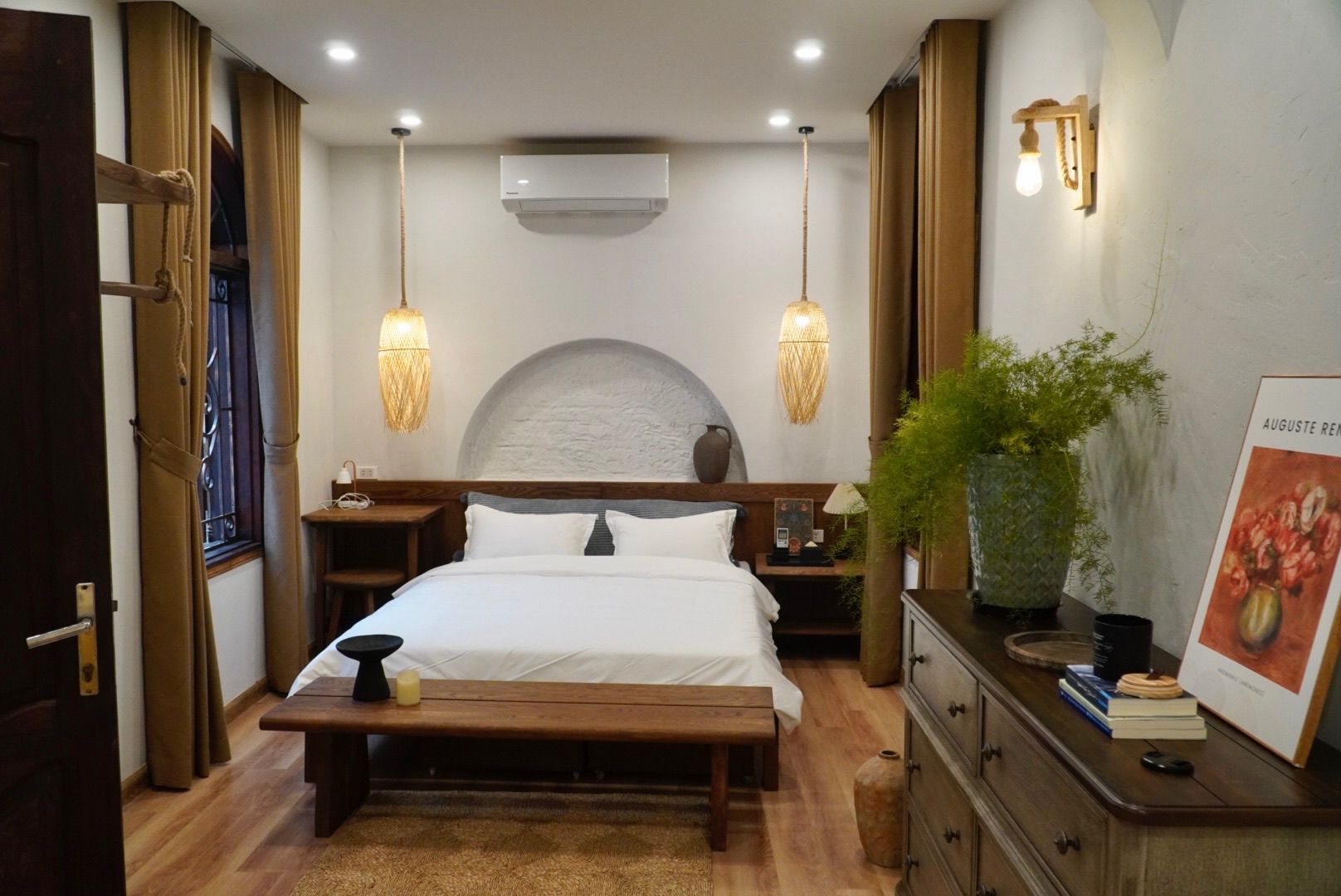 Un dormitorio con cama, mesitas de noche, un banco, cómoda, planta en maceta y aire acondicionado de pared.