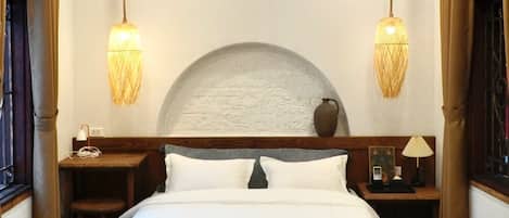 A hotel room with a large bed, bedside tables, a bench, an air conditioning unit, and two hanging lamps.