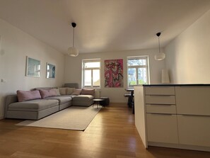 A modern living room with a grey sectional sofa, a small round table, and a kitchen area with white cabinets. - Modern City Apartment at Sendlinger Tor – 5 minutes from downtown – right in the heart of Munich (München)