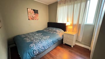 Ein Schlafzimmer mit einem Bett, einem Nachttisch, einer Lampe, einem Gemälde und einem Fenster mit Vorhängen.