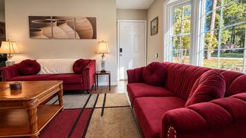 Ein Wohnzimmer mit zwei roten Sofas, einem Holztisch und einem Teppich. An der Wand hängt ein Gemälde und durch das Fenster ist ein Blick auf Bäume möglich.