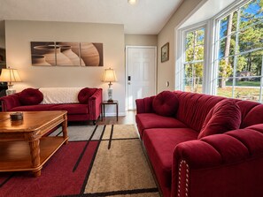 A living room with two red sofas, a wooden coffee table, and a rug. There is a painting on the wall and a window with a view of trees. - Cozy 3-bedroom Rocky Mount Home Retreat With Full Kitchen & Fenced Yard (Rocky Mount)