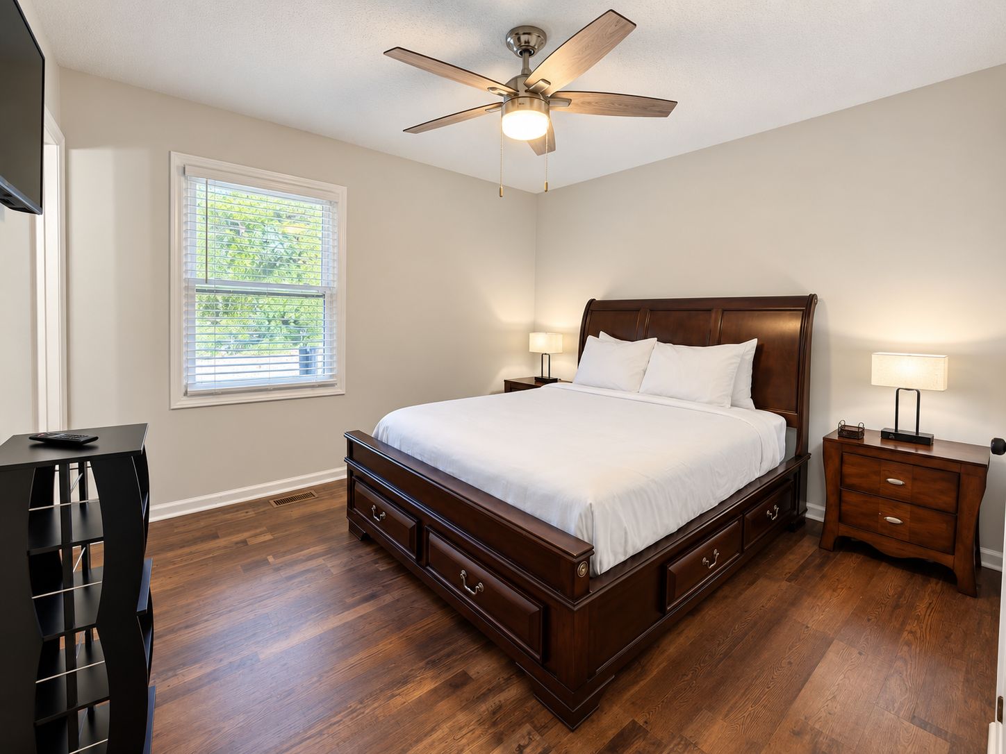 A bedroom with a large bed, wooden headboard, bedside tables with lamps, a ceiling fan, and a window with blinds.
