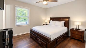 A bedroom with a large bed, wooden headboard, bedside tables with lamps, a ceiling fan, and a window with blinds.