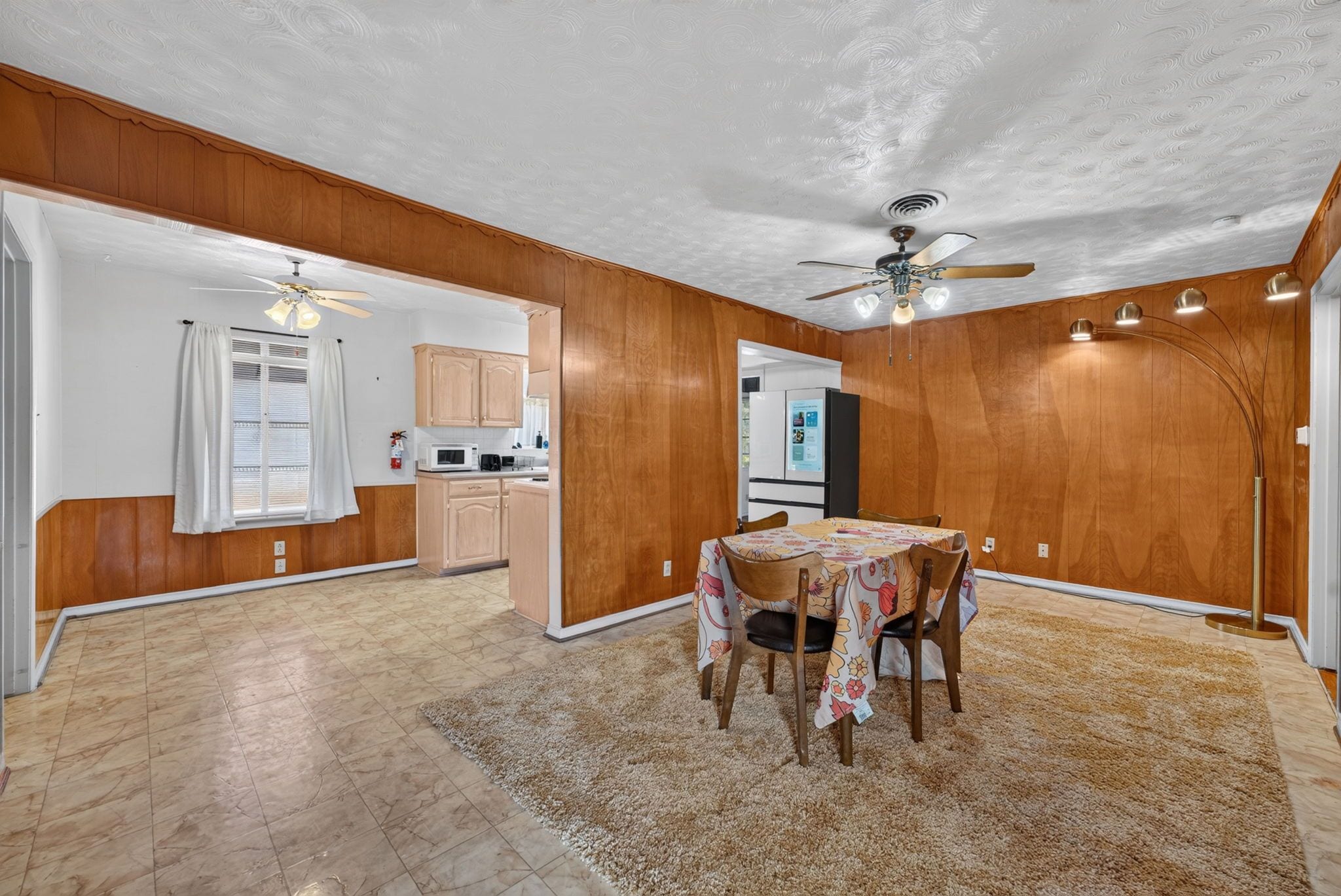 A spacious room with a dining table, chairs, a ceiling fan, and a kitchen area with cabinets and appliances.