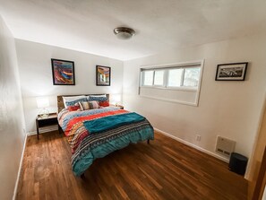A bedroom with a wooden floor, a bed with a colorful blanket, a nightstand with a lamp, and a window with a view. - Cozy midcentury home in South Eugene - 1 mi. to UO + walkable to cafes & trails (Eugene)