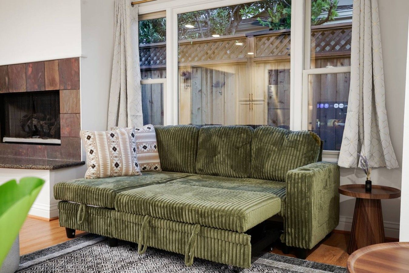 A green sofa with a fireplace, a window with curtains, and a small round table with a vase.