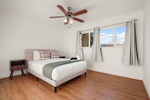 Un dormitorio con cama, mesita de noche, ventilador de techo y ventana con cortinas.