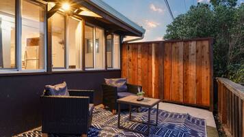 Un patio al aire libre con mobiliario de mimbre y un tapete estampado, una valla de madera y un edificio con grandes ventanales.