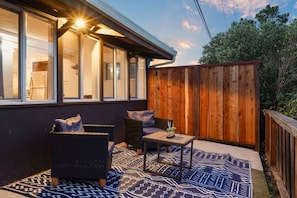 Un patio al aire libre con mobiliario de mimbre y un tapete estampado, una valla de madera y un edificio con grandes ventanales.