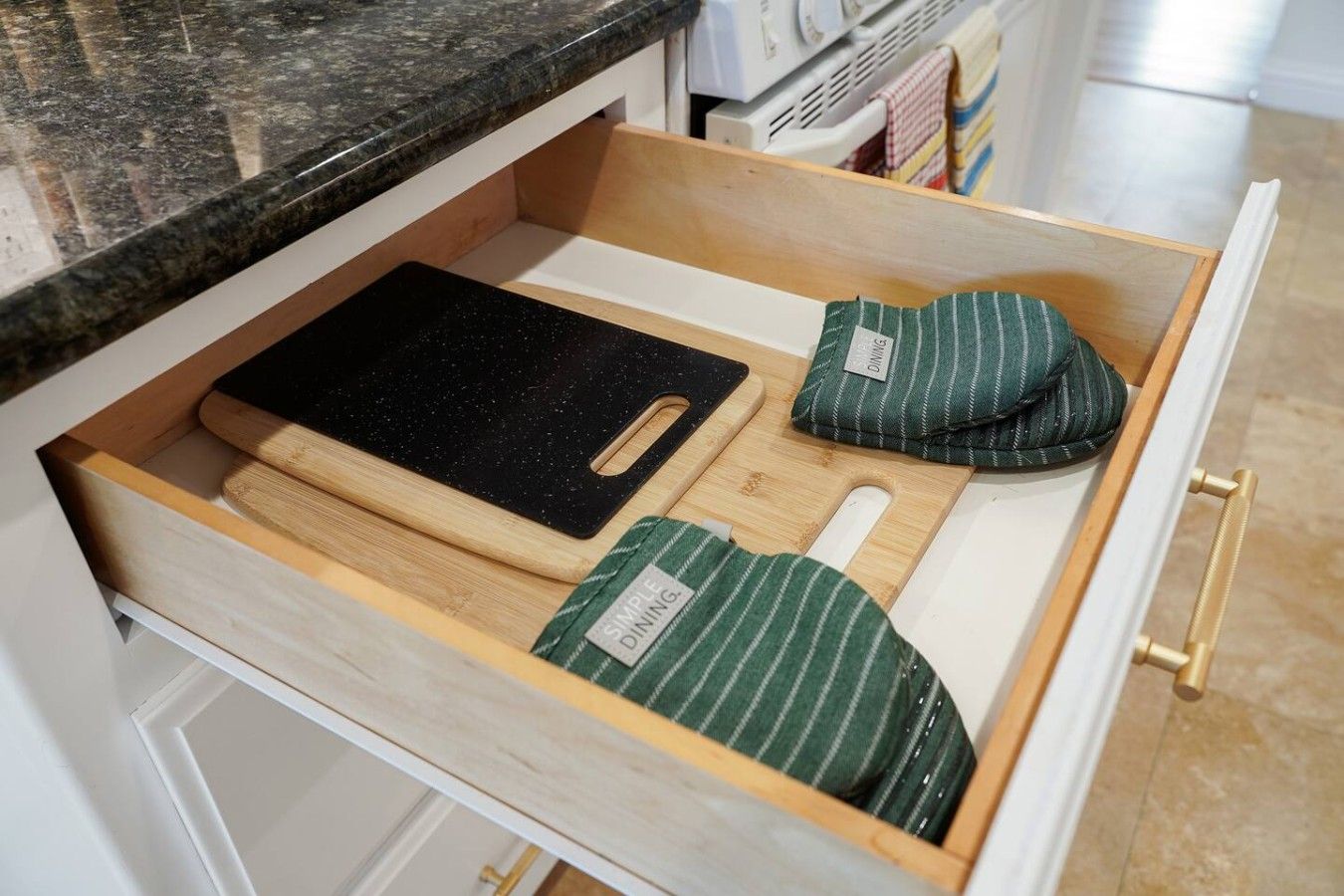 A drawer with a black cutting board, a wooden handle, and two green-striped oven mitts.
