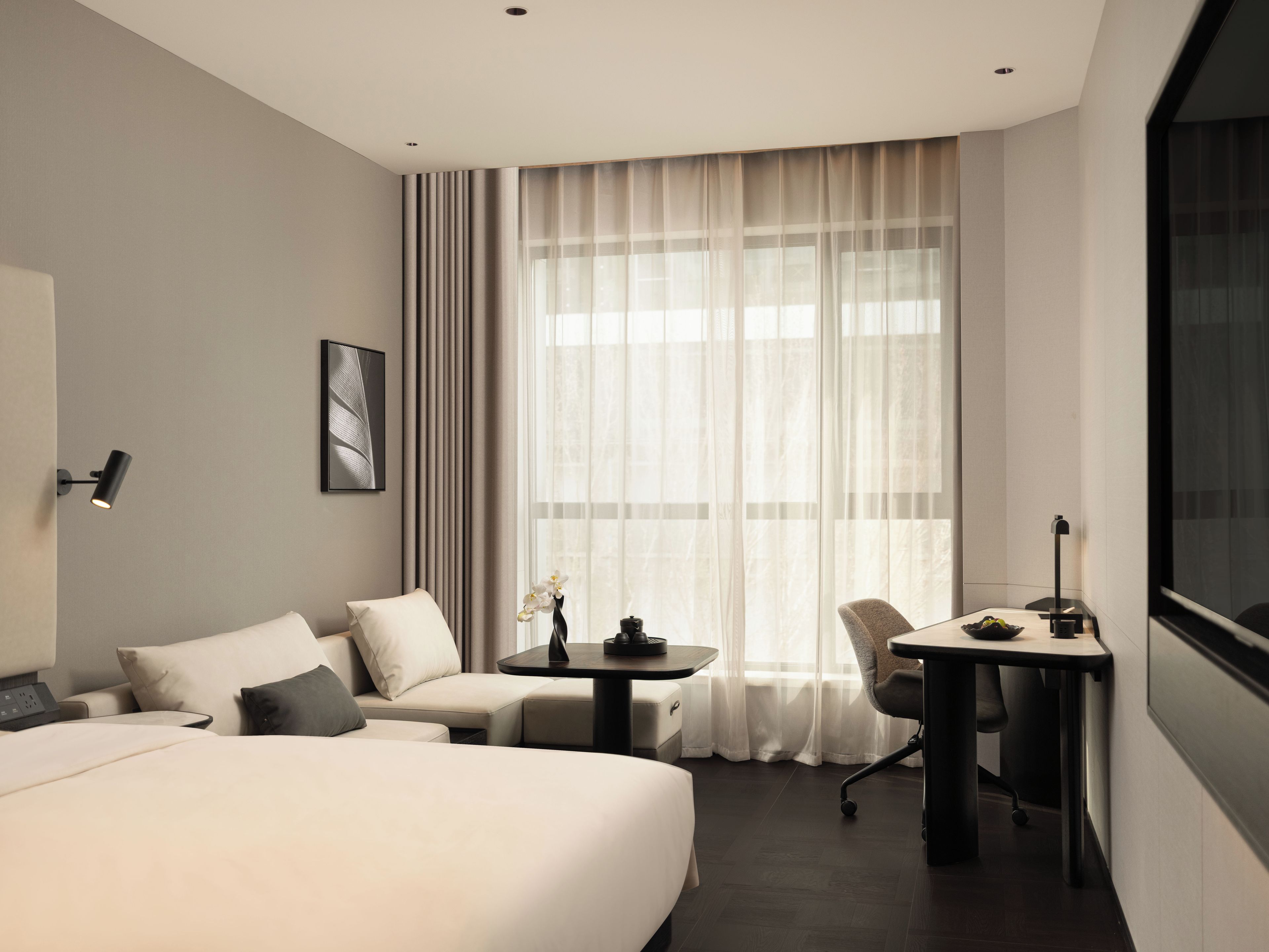 A hotel room with a bed, a sofa, a desk, and a chair. There is a window with curtains, a wall-mounted lamp, and a framed picture.