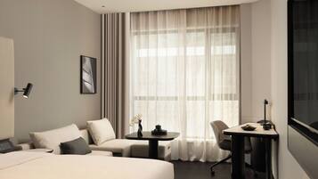 A hotel room with a bed, a sofa, a desk, and a chair. There is a window with curtains, a wall-mounted lamp, and a framed picture.