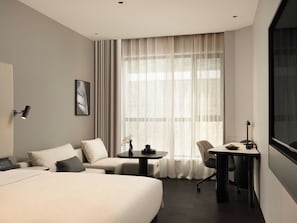 A hotel room with a bed, a sofa, a desk, and a chair. There is a window with curtains, a wall-mounted lamp, and a framed picture. - ShenyangStation West Square Atour Hotel (Shenyang)