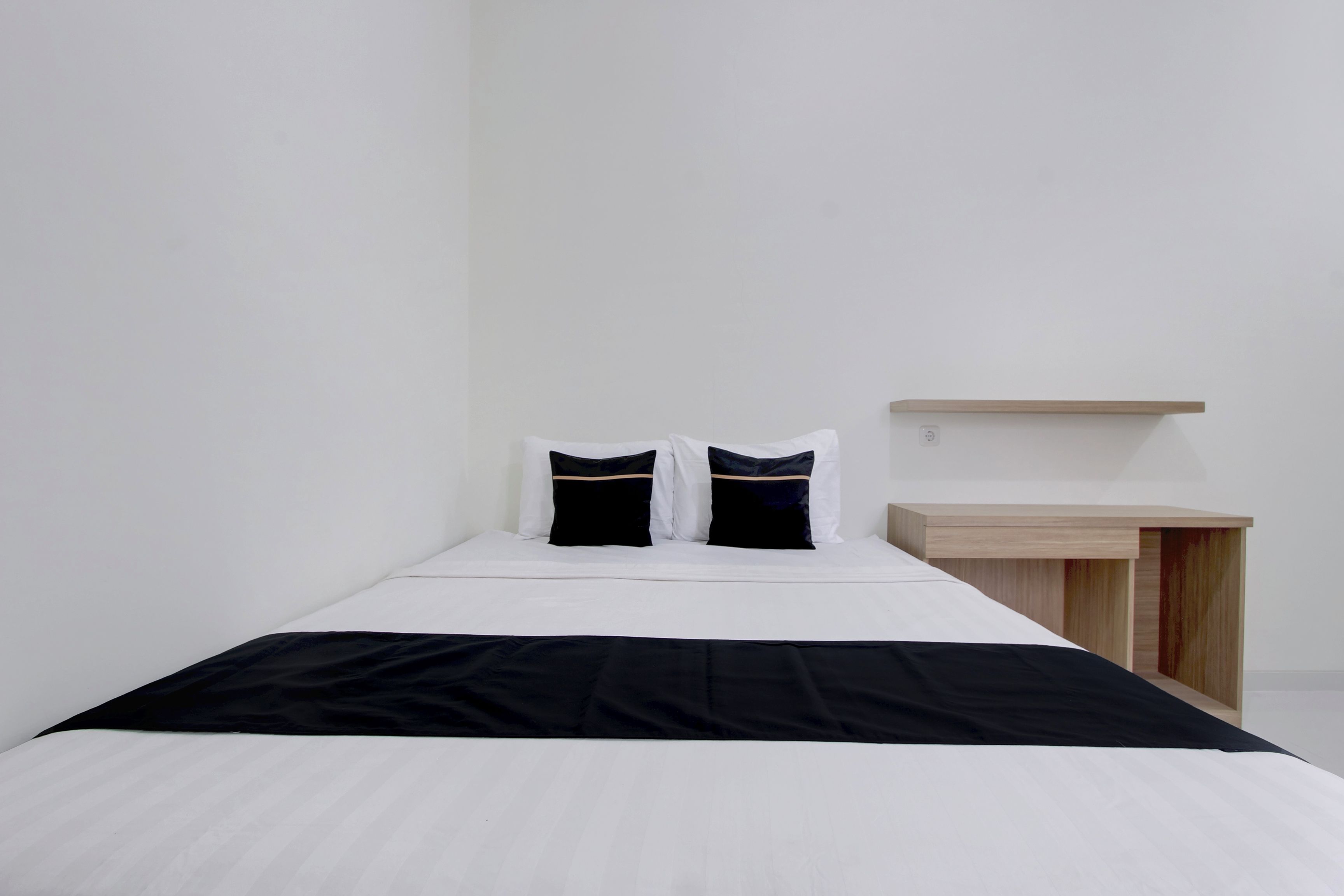 Una cama individual con colcha blanco y negro, dos almohadas negras y una mesita de noche de madera con repisa de color claro.