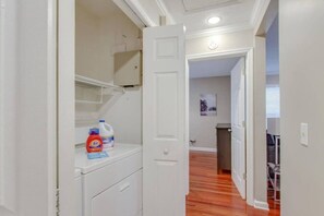 A hallway with a white door, a shelf with cleaning supplies, and a view into another room with a wooden floor. - Stylish Getaway Prime Location Near Downtown (Nashville)