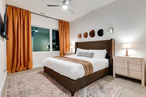Un dormitorio con cama, mesita de noche, lámpara, ventilador de techo y ventana con cortinas.