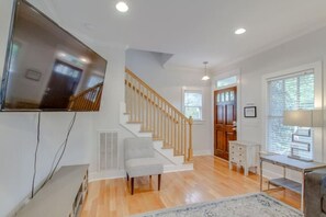 A spacious living area with a wooden staircase, a flat-screen TV mounted on the wall, a grey armchair, and a desk with a lamp. - 3BR + Gated Parking | Sleeps 8 /Mins to Broadway (Nashville)