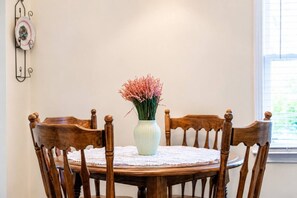 Ein runder Holztisch mit weißer Spitzenleinentischdecke, umgeben von Holzstühlen mit gedrehten Leisten. In der Mitte des Tisches steht eine Vase mit rosa Blumen.