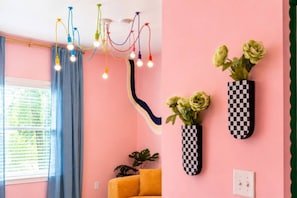 Ein Zimmer mit einer rosafarbenen Wand, einem gelben Sofa, einer karierten Vase mit Blumen und einer mehrfarbigen Pendelleuchte.