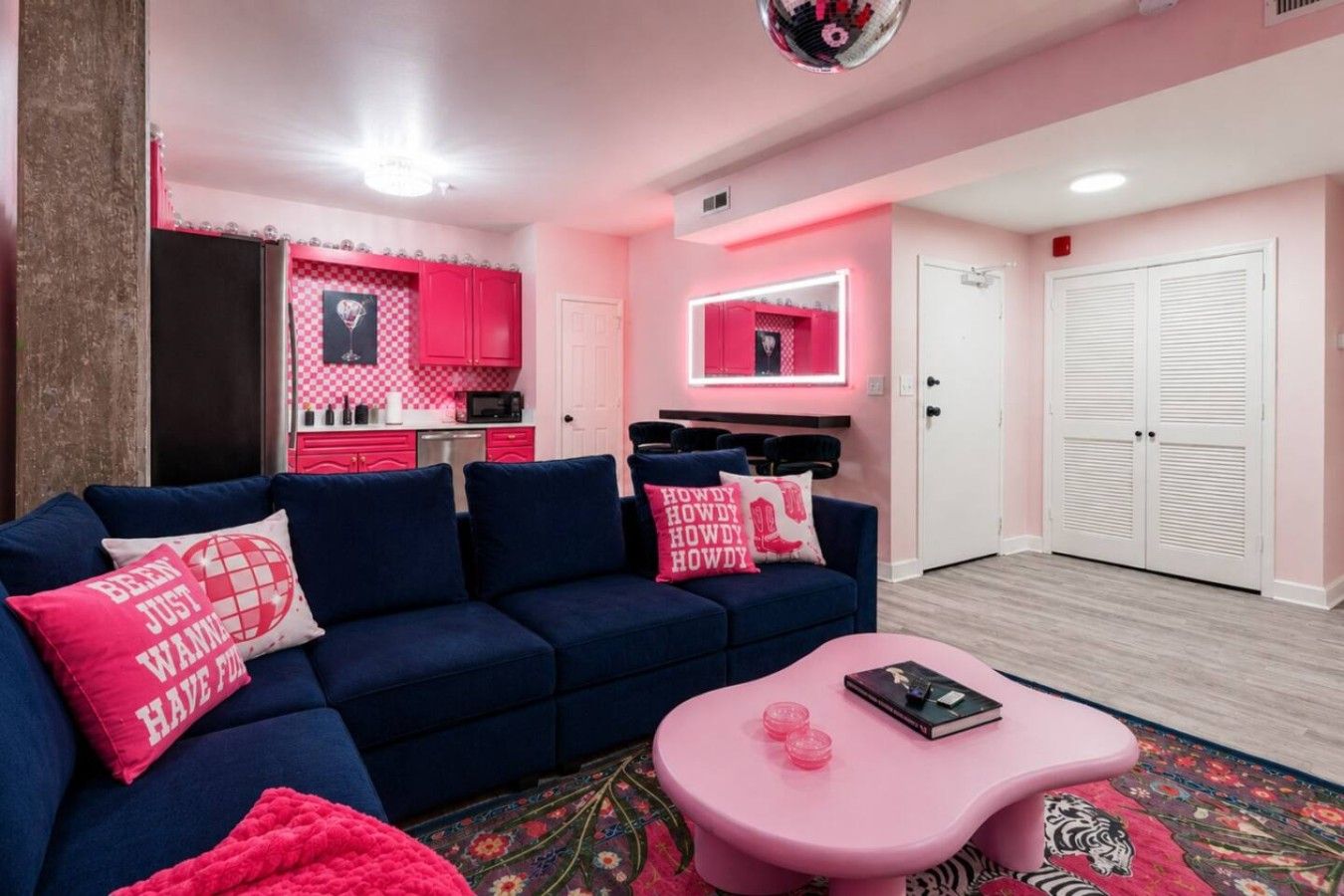 A room with a blue sofa, pink pillows, a pink coffee table, and a kitchen area with pink cabinets and a mirror.