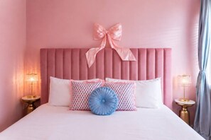 Cama com cabeceira rosa, duas luminárias de cabeceira e almofadas estampadas.