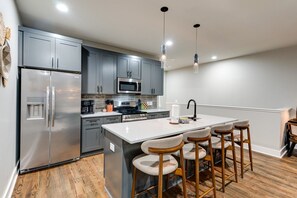 A modern kitchen with a stainless steel refrigerator, a microwave, and a sink. There are bar stools and a large island with a white countertop. - Nashville Stay Near Downtown | 3BR on Fern Ave (Nashville)