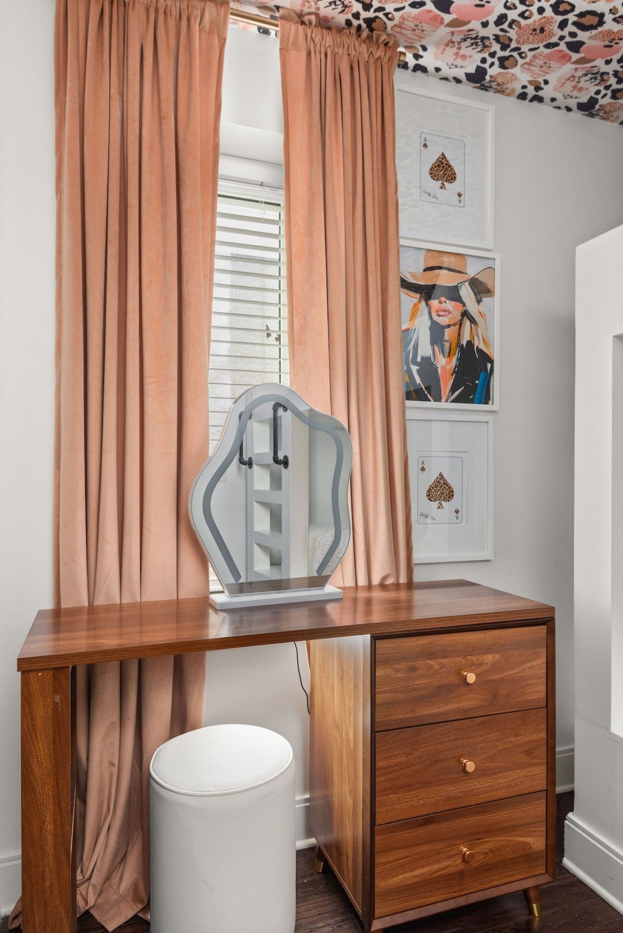 Een houten bureau met een witte prullenbak, een lijst met een speelkaart en een raam met gordijnen.