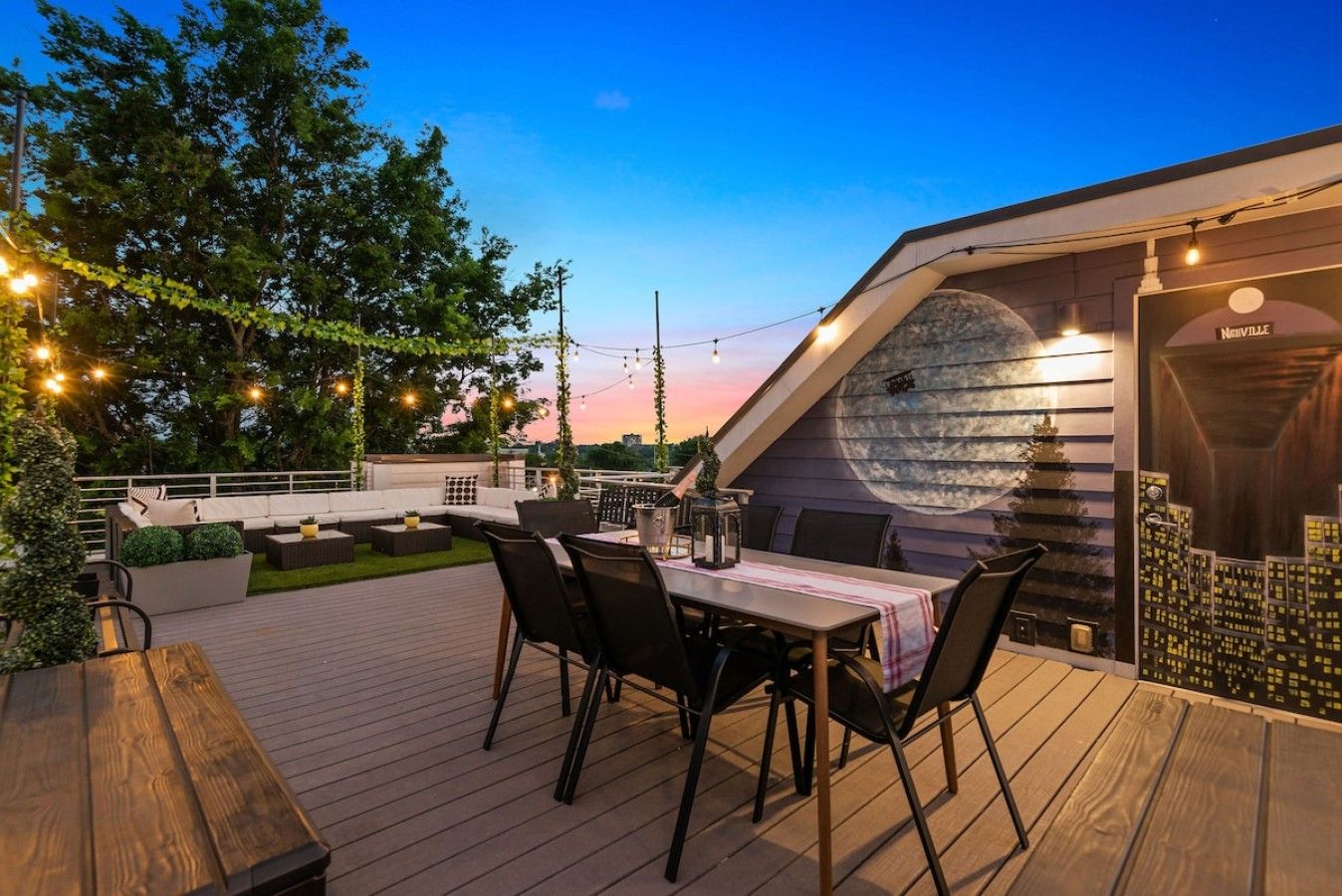 Een terras buiten met een eettafel, stoelen en een bank. Er is een muurschildering op de muur en een sfeerverlichting.