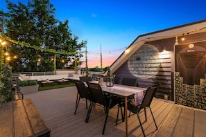 Una terraza al aire libre con mesa de comedor, sillas y sofá. Hay un mural en la pared y luces colgantes.