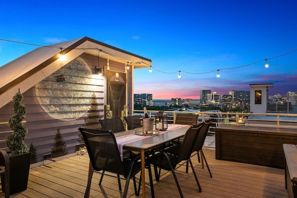 Una terraza en la azotea con mesa, sillas y vistas a la ciudad al anochecer.