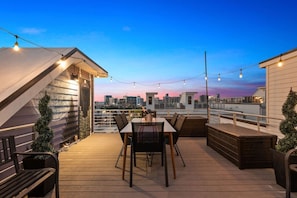 Una terraza en la azotea con mesa y sillas, un muelle de madera y vistas al horizonte de la ciudad al anochecer.