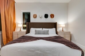 Habitación de hotel con una cama grande, dos mesitas de noche, un ventanal con cortinas y una pared con cuatro sombreros.