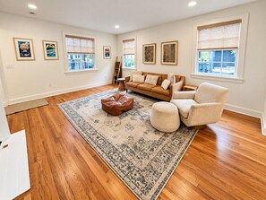 A living room with hardwood flooring, a patterned rug, a brown leather ottoman, a beige sofa, and a white armchair. - Pet Friendly Home Near Vanderbilt & Centennial Park | 6-BR Home (Nashville)