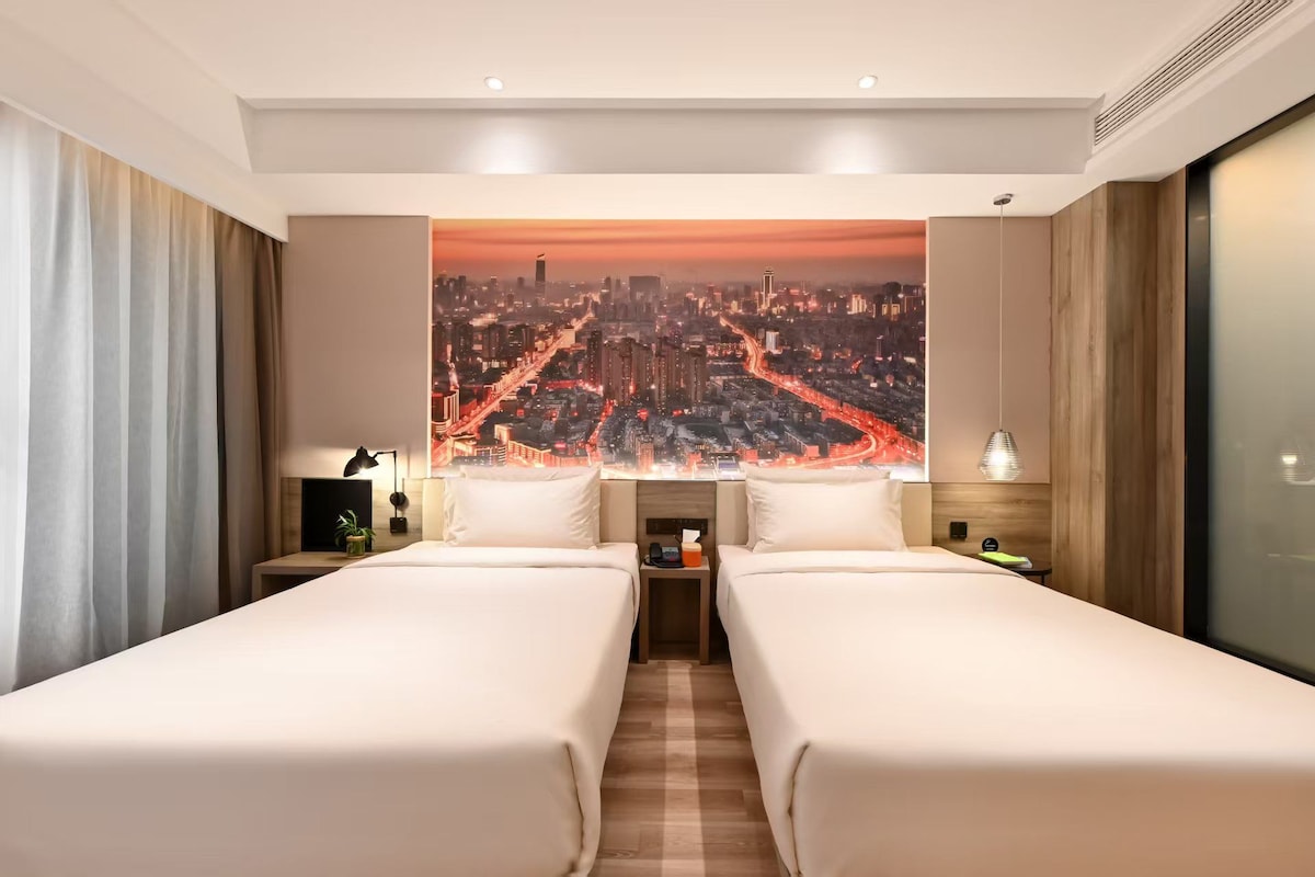 Habitación de hotel con dos camas, vista panorámica de la ciudad, una mesita de noche con lámpara y cortinas en la ventana.