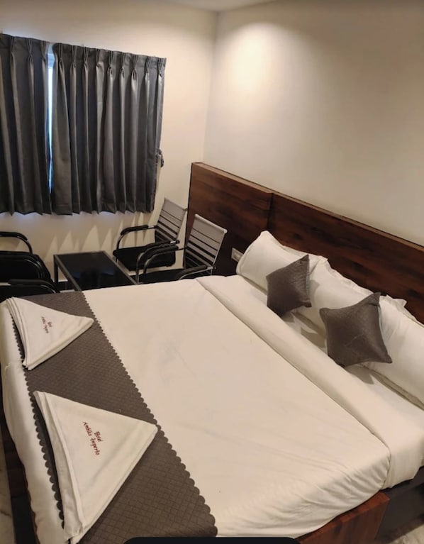 A neatly made bed with pillows and a blanket, a wooden headboard, a window with curtains, and a chair by the window. - Hotel Ambika Imperio (Vijayawada)