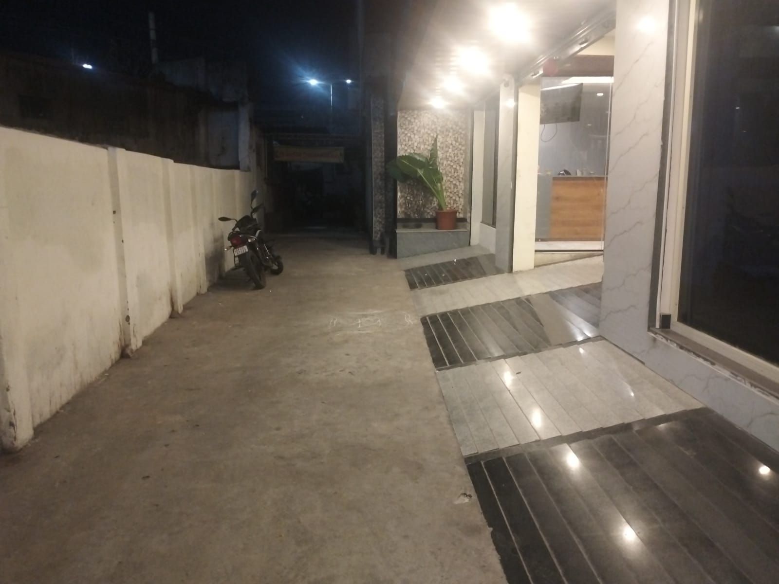A corridor with a motorcycle parked on the left, leading to a reception area with a potted plant and a wooden counter.