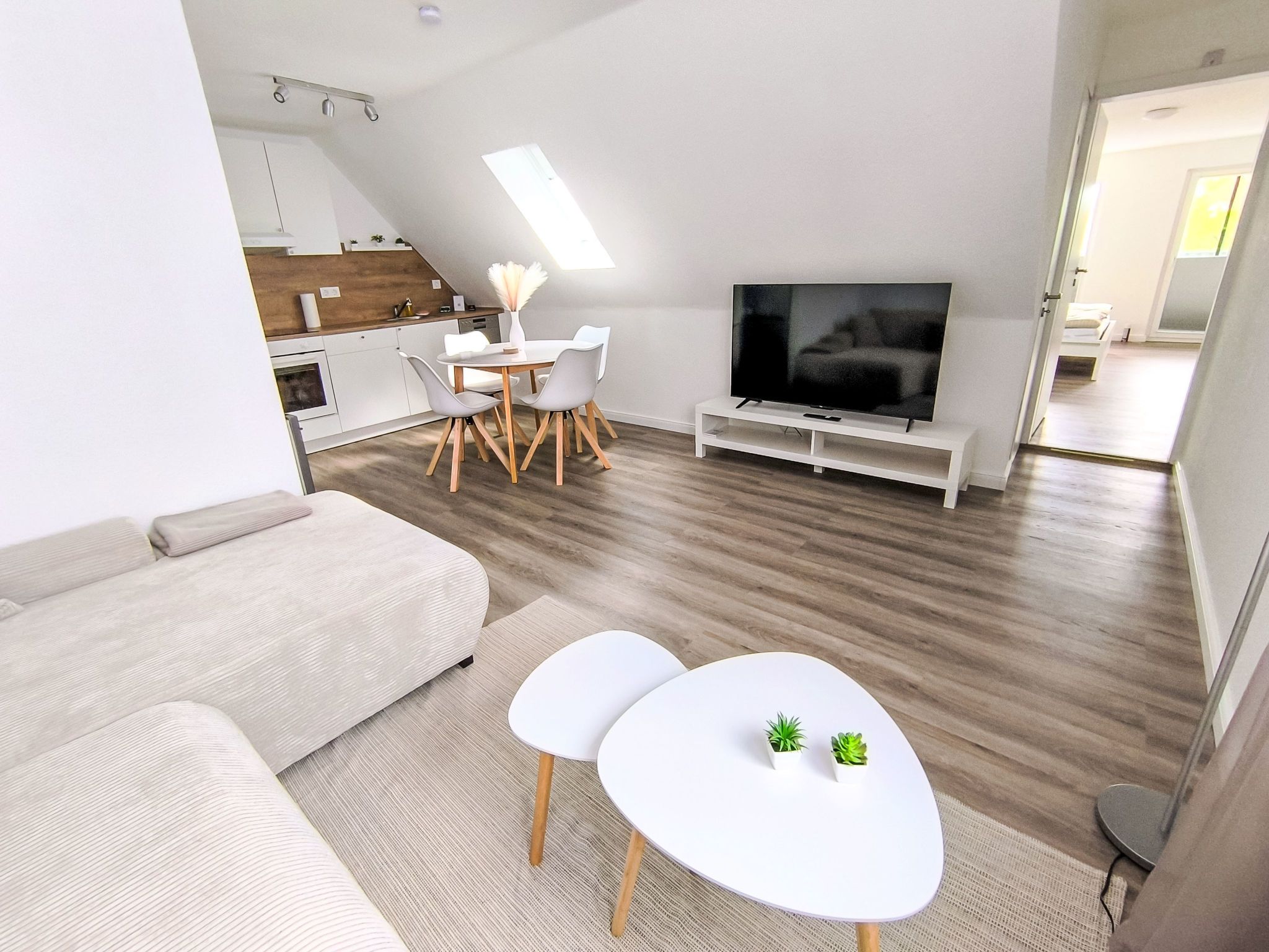A modern living room with a sofa, a dining table with chairs, a flat-screen TV, and a kitchen area in the background.