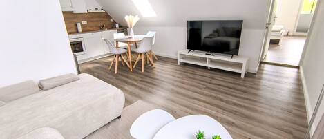 A modern living room with a sofa, a dining table with chairs, a flat-screen TV, and a kitchen area in the background.