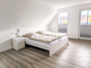 Un dormitorio con techo inclinado, una cama con estructura blanca, una mesita de noche con lámpara y una ventana con persianas.
